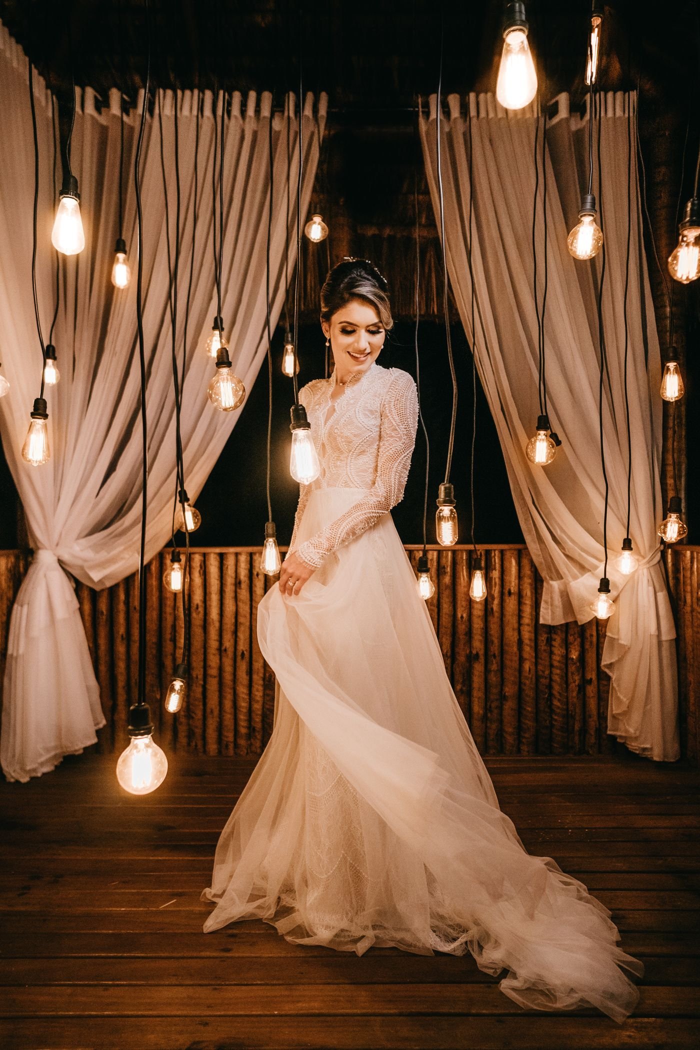 Bride in a lace-bodice gown with a long tulle train under hanging bulb lights.